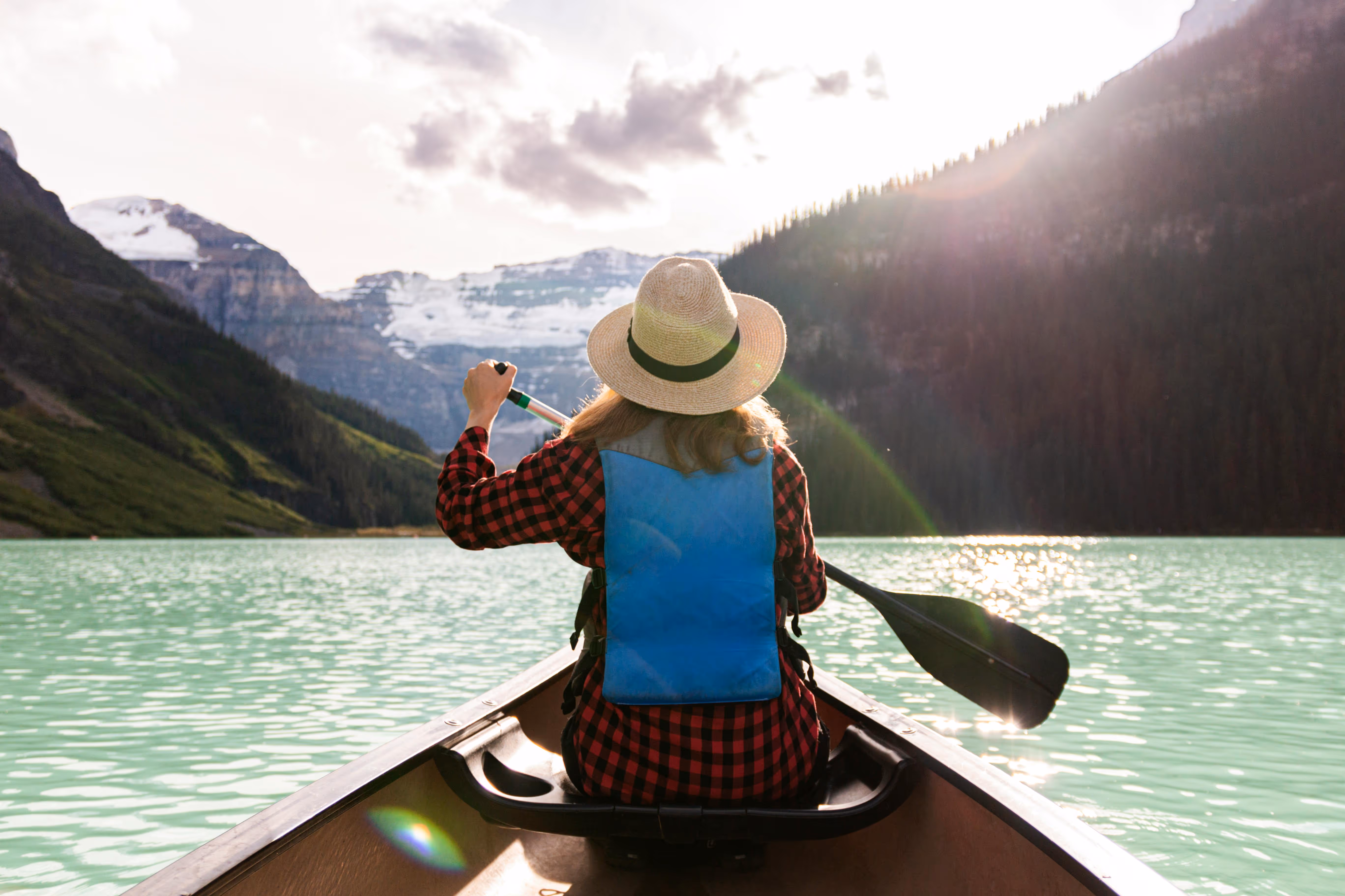 Back view of a woman kayaking on a calm lake with mountains around and the bright sun ahead