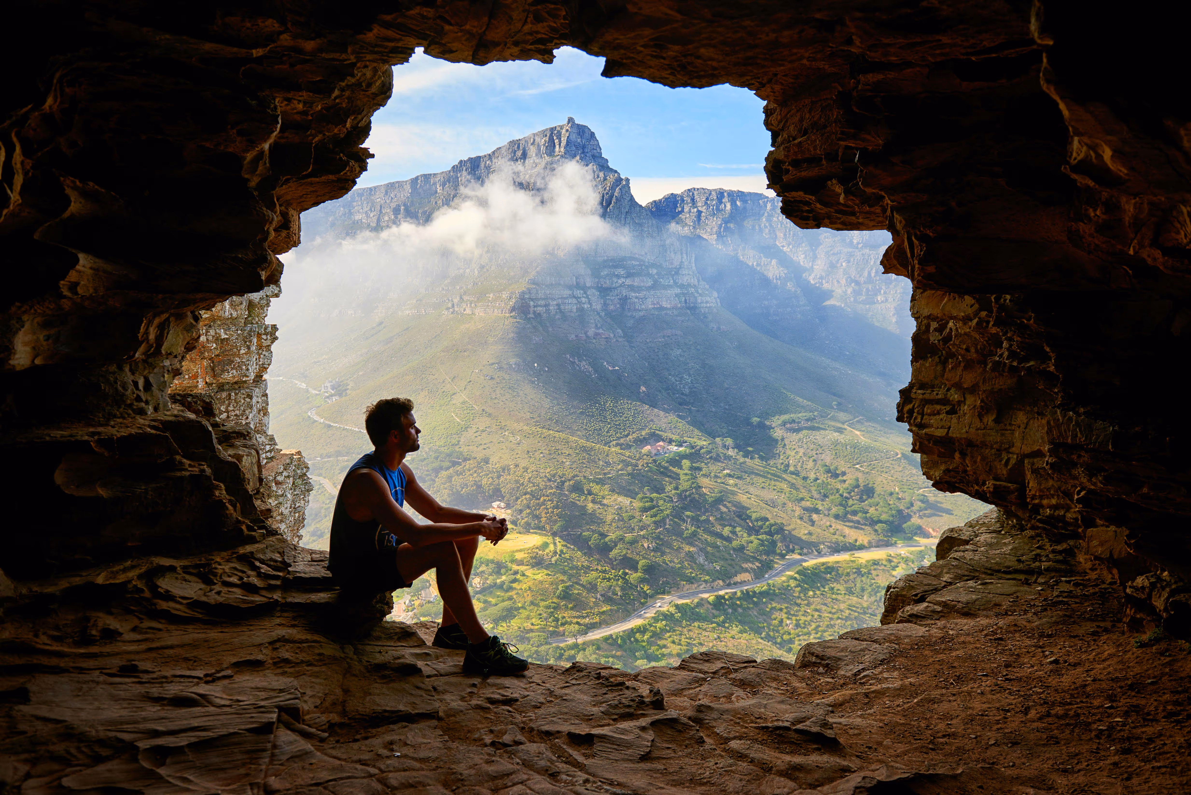 View from inside a cave showing a man sitting at its edge, with mountains and nature visible outside